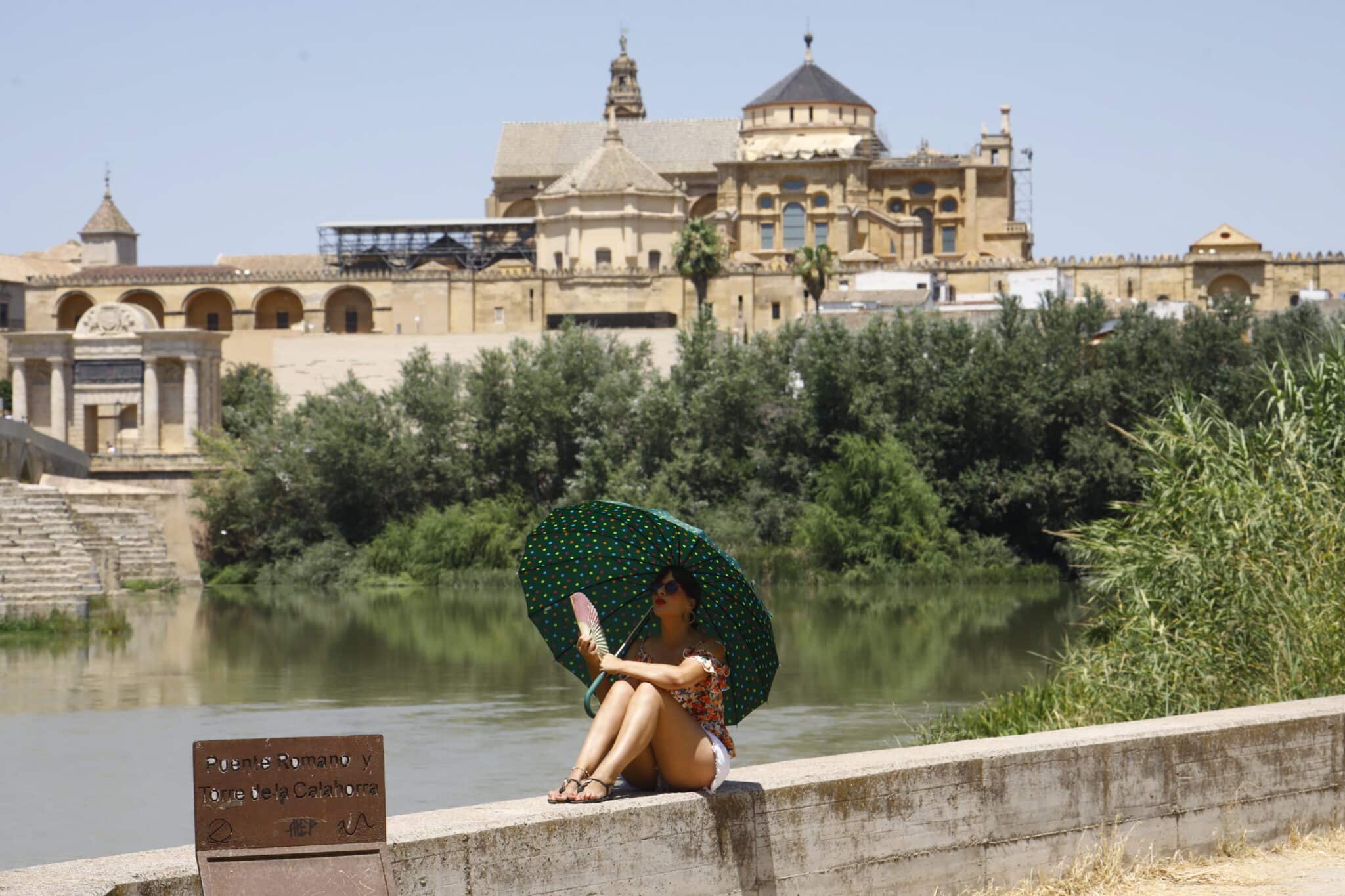 Una turista se protege del calor con un paraguas y un abanico a la orilla del río Guadalquivir cerca de la Mezquita-Catedral de Córdoba ( al fondo) hoy lunes cuando la Agencia Estatal de Meteorología (Aemet),