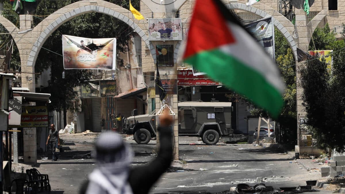 Un palestino sostiene una bandera palestina frente a vehículos militares israelíes en los accesos al campamento de refugiados de Yenín.