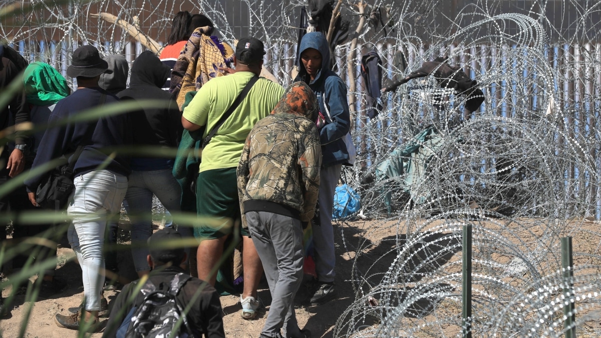 Migrantes esperan entre los alambres de púas cerca del muro que separa la frontera estadounidense